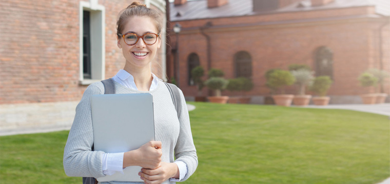 Eine junge Frau mit Laptop im Arm steht lächelnd auf dem Campus.