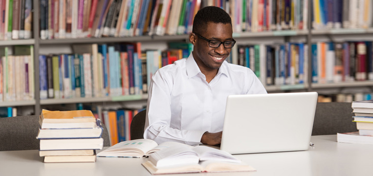Ein Student sitzt zwischen Bücherstapeln lächelnd vor seinem Laptop.