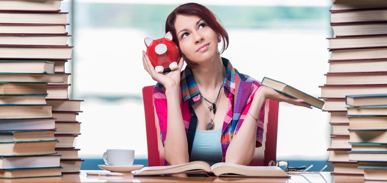 Junge Frau sitzt mit Sparschwein in der Hand zwischen Stapeln von Büchern.